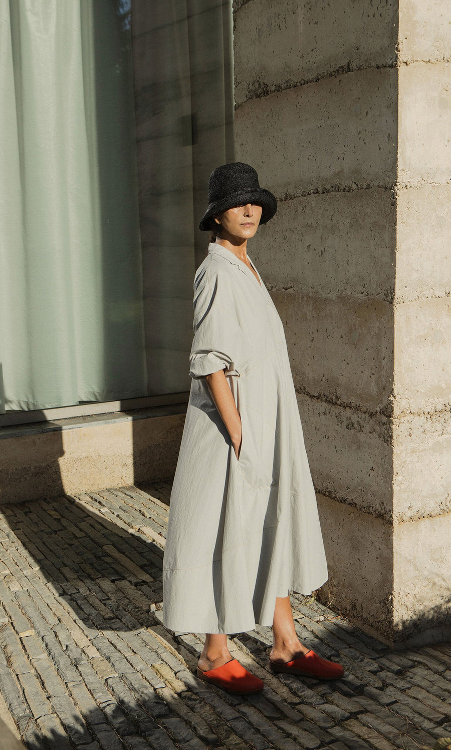 Plümo- Person in a loose light gray dress, Straw Bucket Hat, and red shoes stands on a stone path by a modern concrete building.