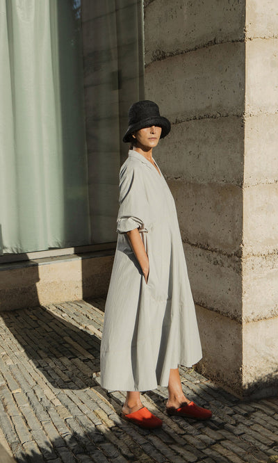 Plümo- Person in a loose Kaisa Dress with balloon sleeves, black hat, and red shoes stands by a concrete building in sunlight.