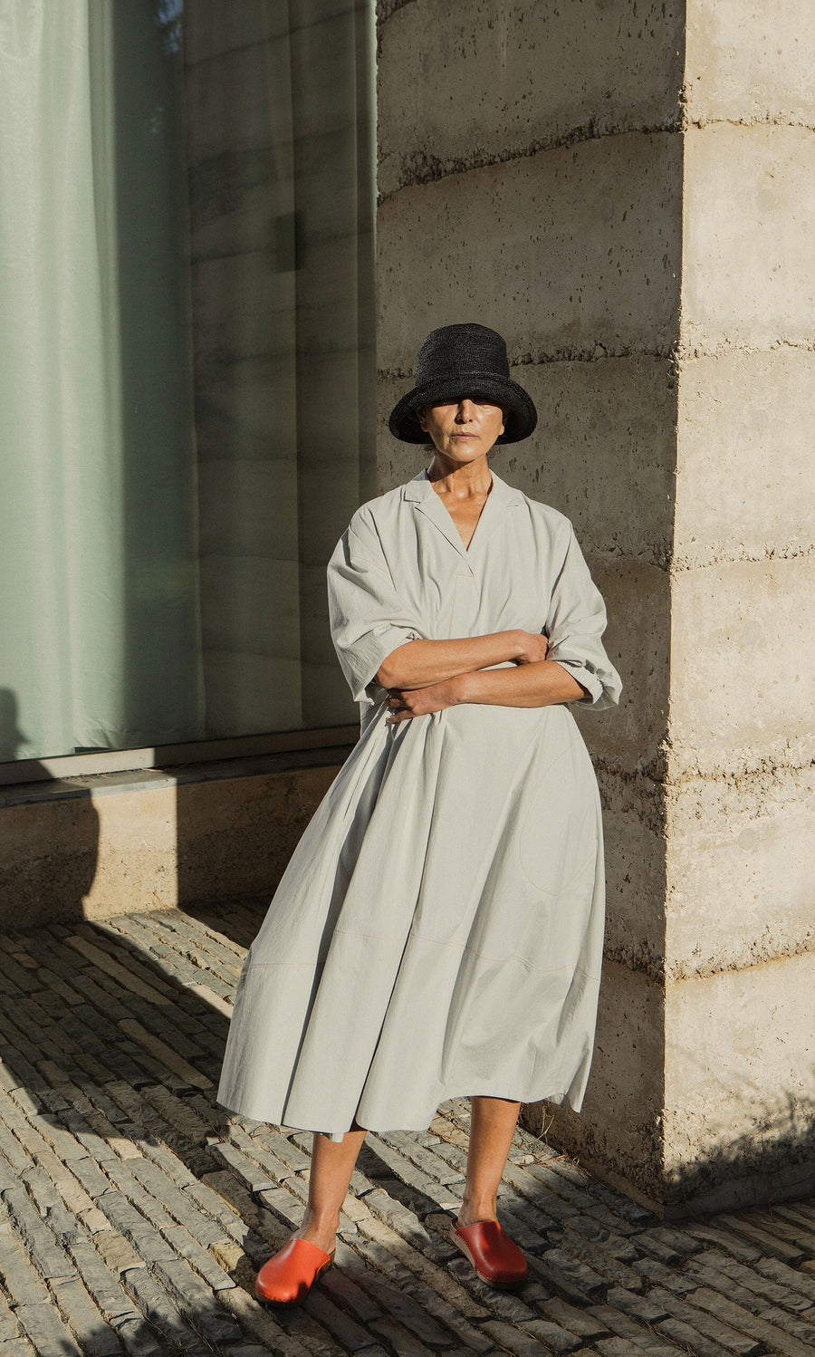 Plümo- Woman in a loose light gray dress and Straw Bucket Hat stands by a stone wall in sunlight, wearing red shoes.