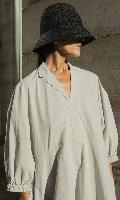 Plümo- Person wears the Kaisa Dress with balloon sleeves and a black woven hat, standing against a sunlit concrete wall.