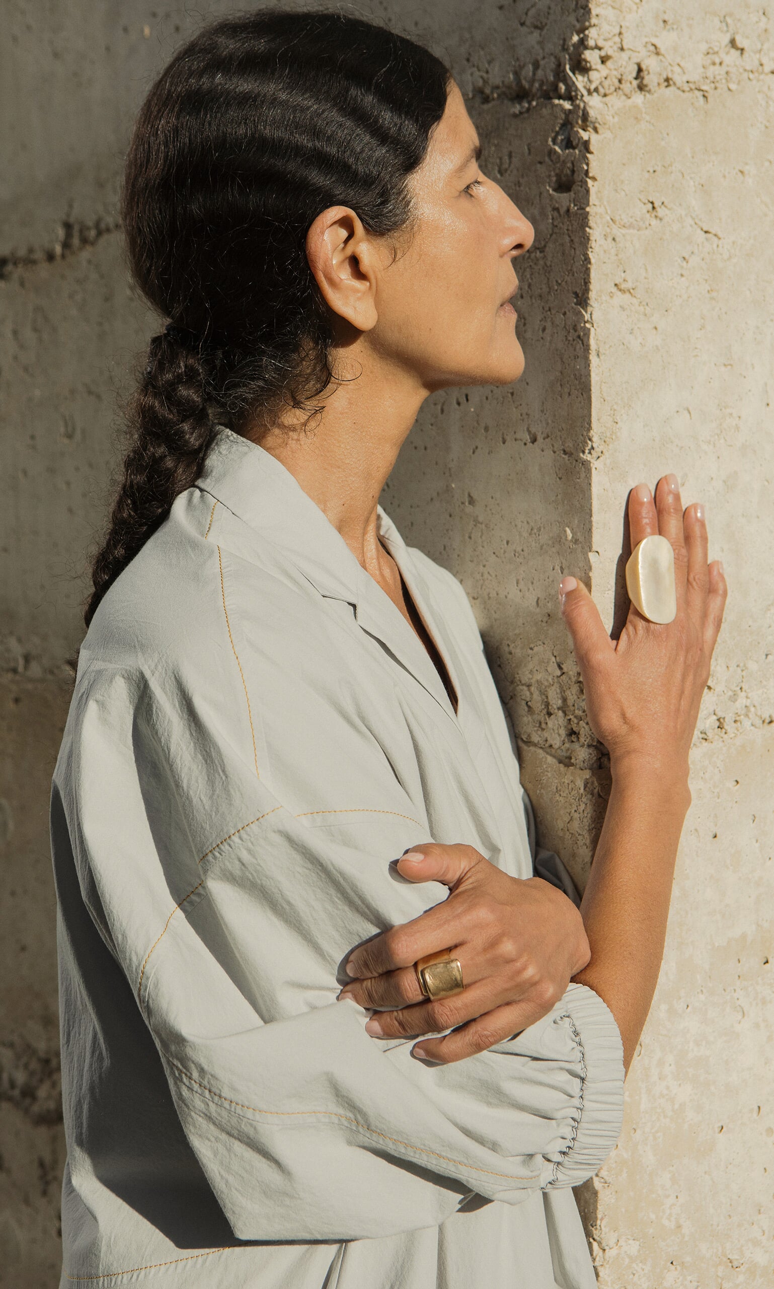 Plümo- A woman with dark braided hair in a light shirt stands sideways, her hand wearing the Delta Ring touches the rough wall.