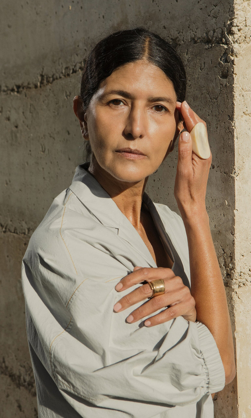 Plümo- Woman with dark hair in a bun, wearing light clothes and the Delta Ring, stands by a concrete wall, touching her face.