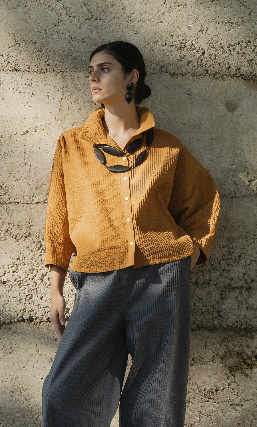 Plümo- A woman in a mustard yellow shirt and gray pants wears the Benin Necklace, standing by a textured wall.