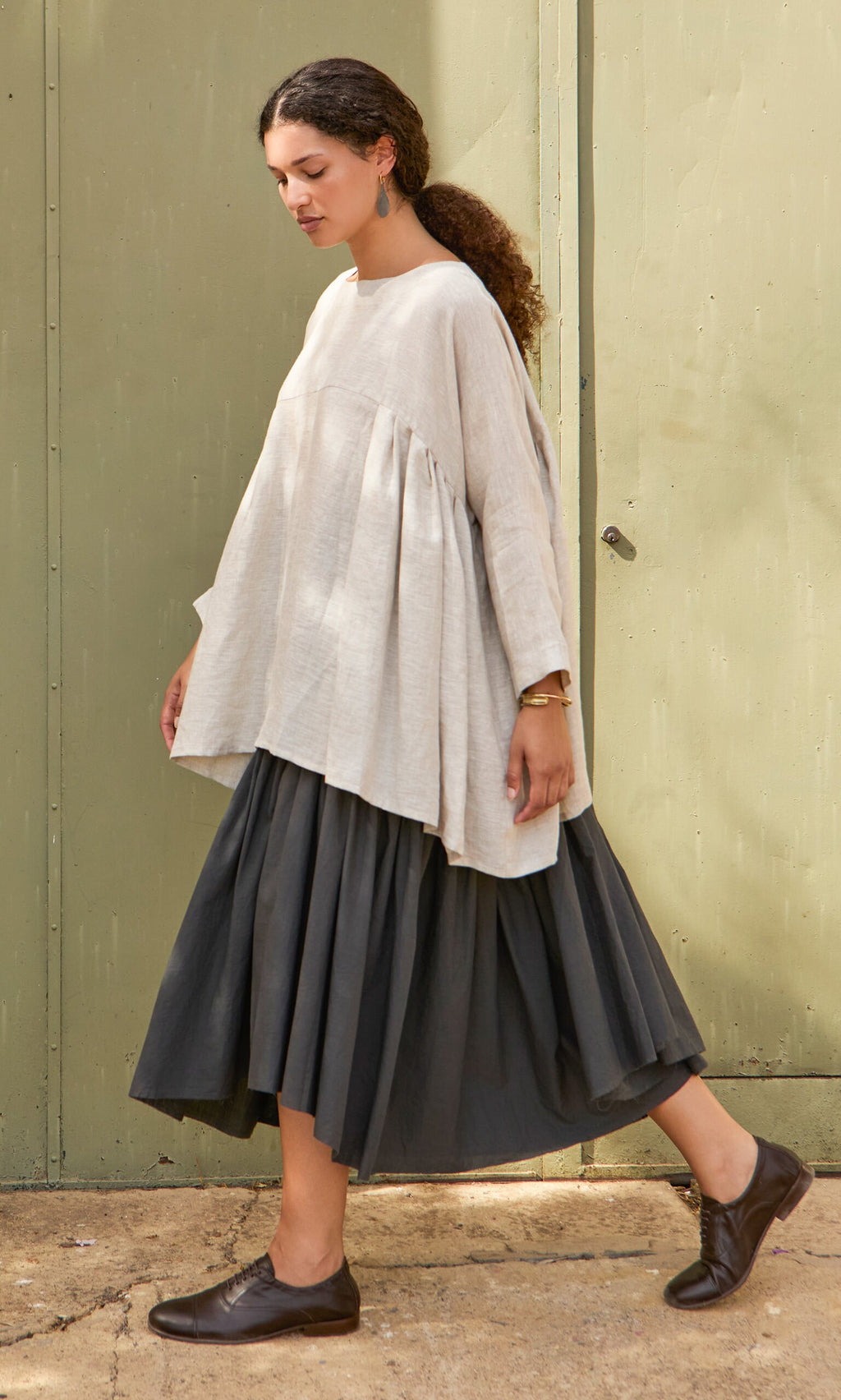 Plümo- Woman in a Kelli Blouse, black skirt, and black shoes walks by a light green door on a sunny day.