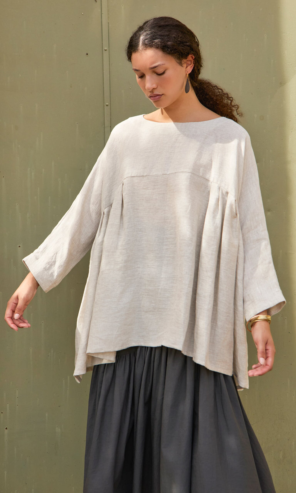 Plümo- Woman in the Kelli Blouse and a dark, flowing skirt stands against a muted green wall.