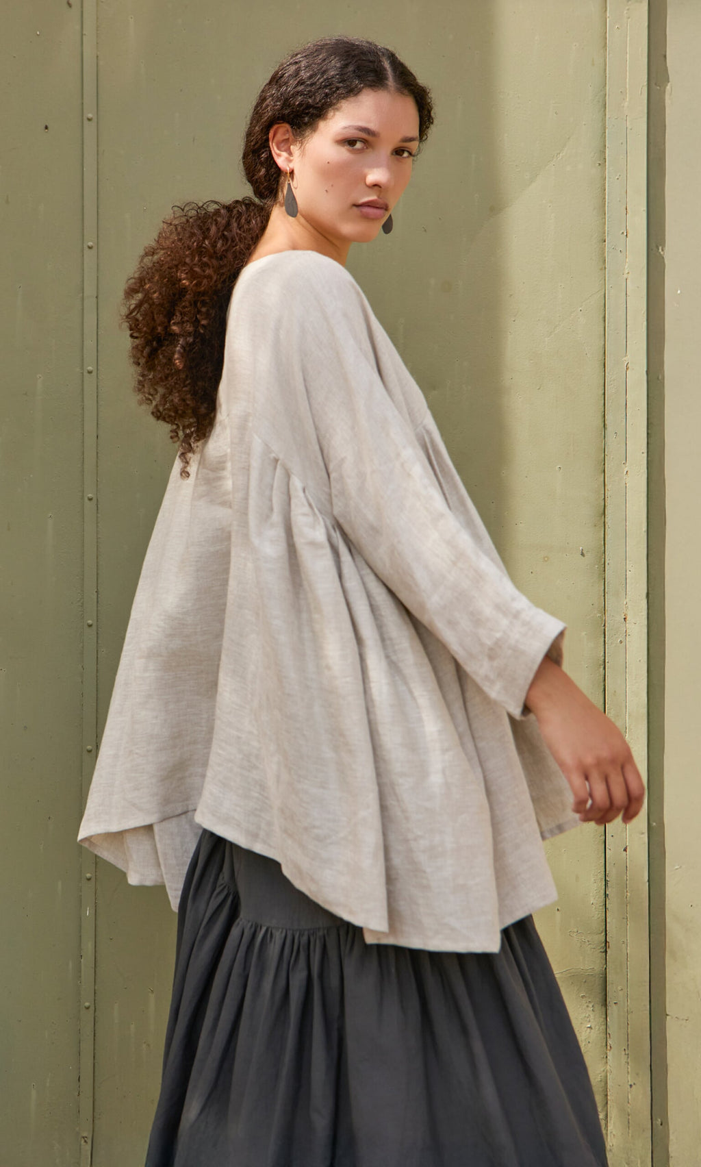 Plümo- Woman with curly hair wears the Kelli Blouse and a long dark skirt, standing sideways against a green wall.