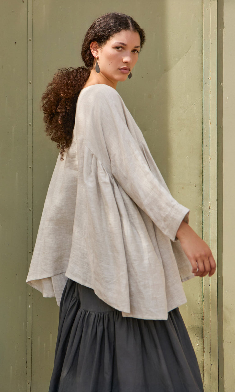 Plümo- Woman with curly hair wears the Kelli Blouse and a long dark skirt, standing sideways against a green wall.