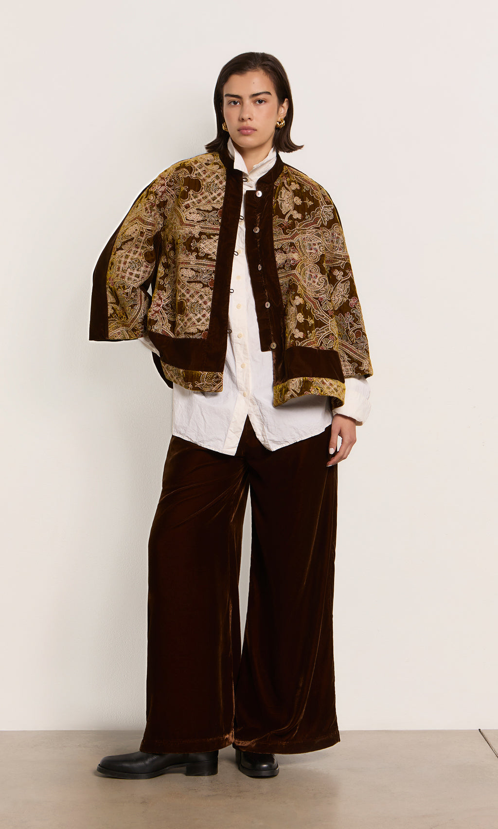 Plümo  Person in maroon velvet pants, white shirt, and patterned cape stands by a plain wall.