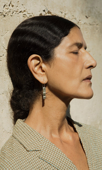 Plümo- Woman with dark wavy hair leans against a sunlit wall, wearing a checked jacket and the Kenan Earrings.