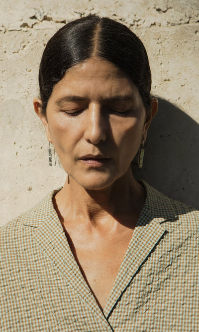 Plümo- Woman with dark hair and closed eyes stands against a textured wall, wearing checkered clothing and Kenan Earrings.