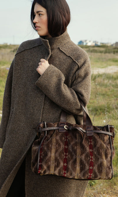 Plümo- Woman outdoors in a sculptural Babette Coat, holding a patterned brown and red handbag with leather straps.
