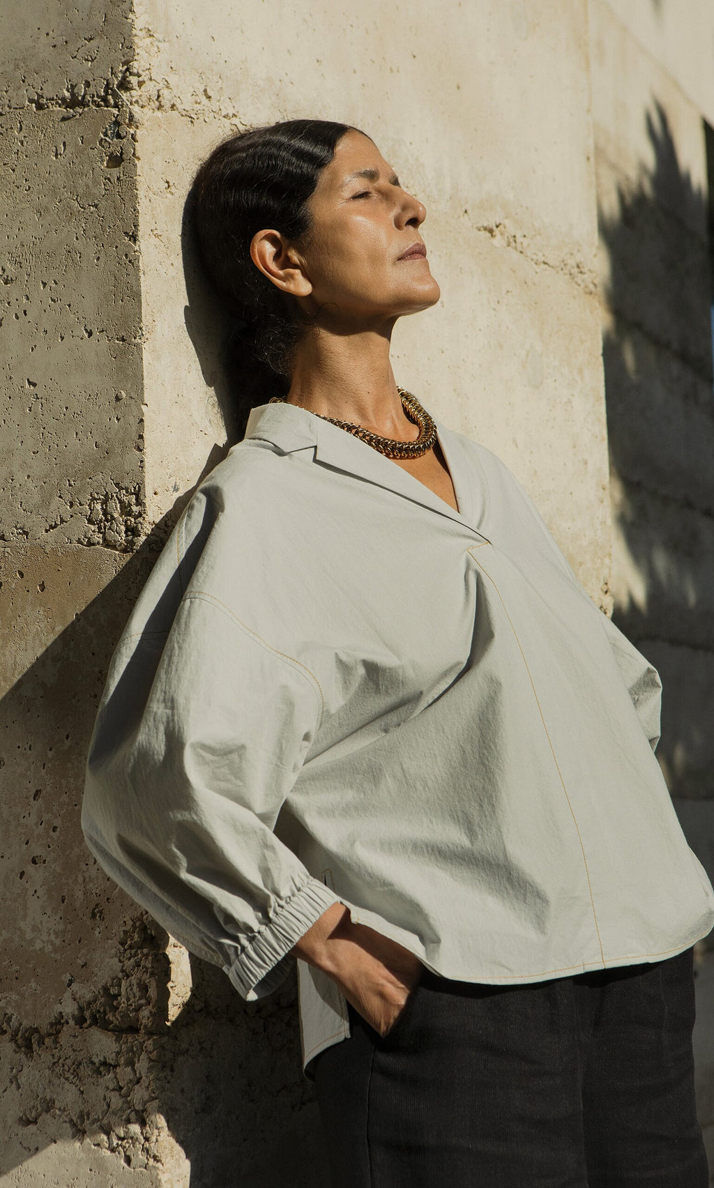 Plümo- Woman in the Fern Blouse - Sky with sculptural sleeves stands against a concrete wall, eyes closed in sunlight.