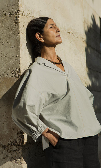 Plümo- Woman in the Fern Blouse - Sky with sculptural sleeves stands against a concrete wall, eyes closed in sunlight.