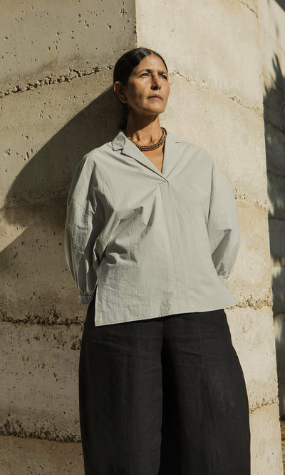 Plümo- A woman in the Fern Blouse - Sky stands against a sunlit wall, gazing thoughtfully into the distance.