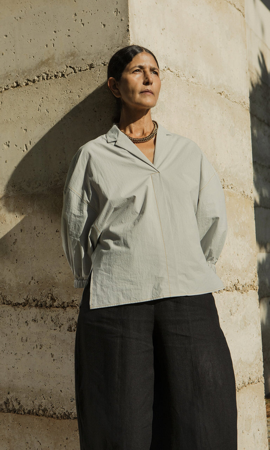 Plümo- A woman in the Fern Blouse - Sky stands against a sunlit wall, gazing thoughtfully into the distance.