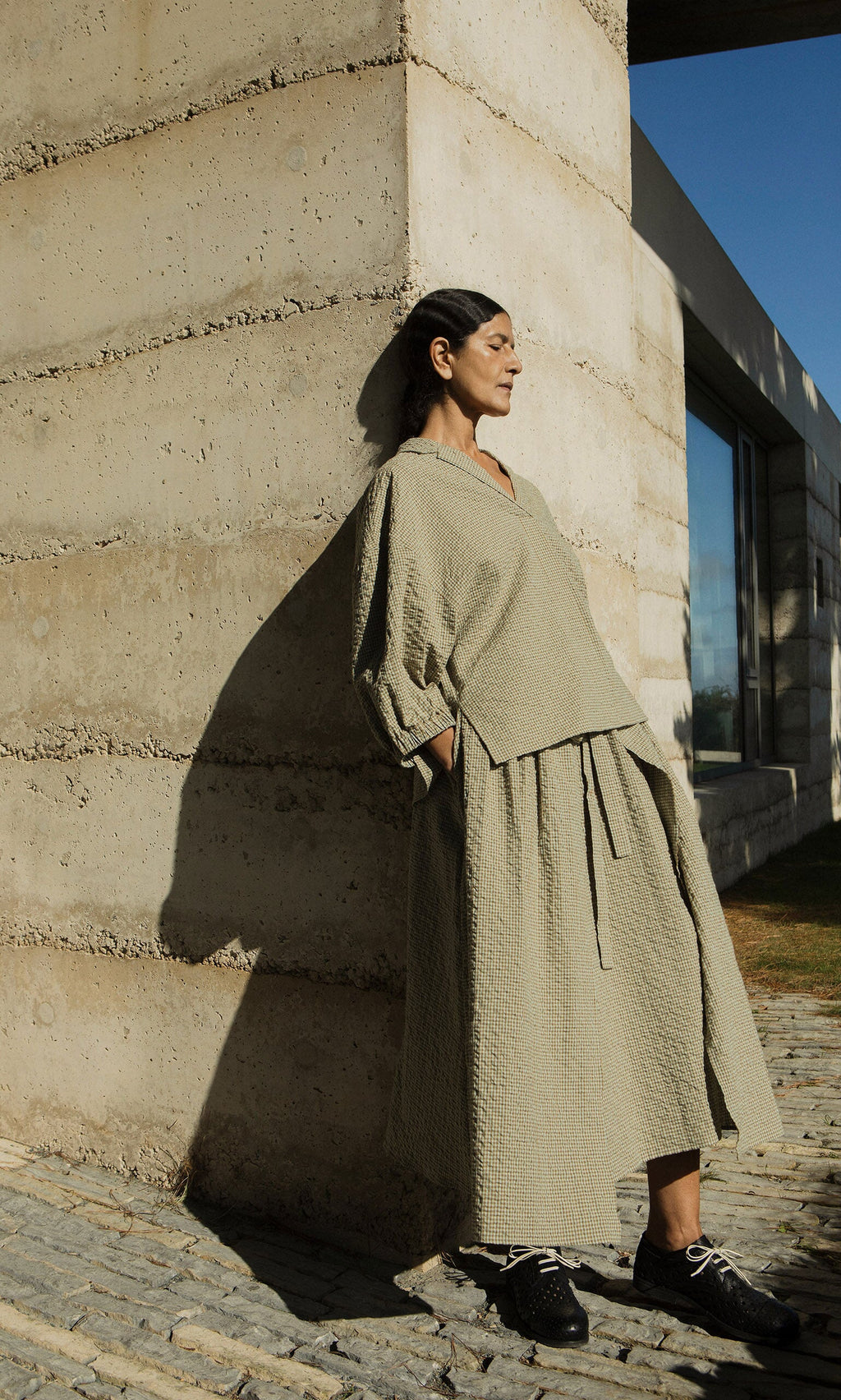 Plümo- A woman in the Fern Blouse leans against a textured concrete wall, casting a shadow in the sunlight.