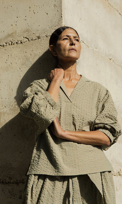 Plümo- Woman in the Fern Blouse stands against a concrete wall, looking up with sunlight on her face and arms crossed.