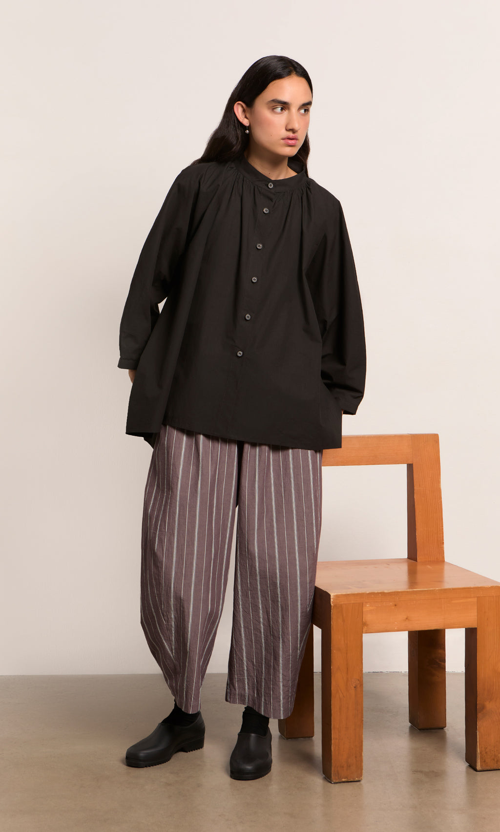 Plümo  Person in a Kroyer Blouse and striped pants stands by a wooden chair against a plain background.