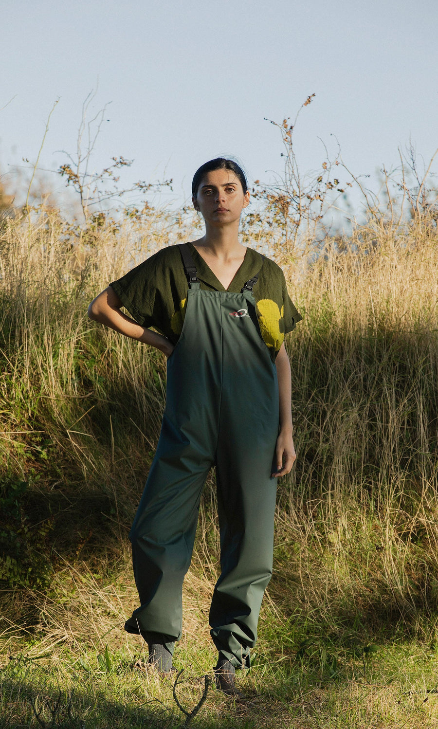 Plümo- Person outdoors in grass, wearing Land Dungarees—green weatherproof with yellow patches—looking at camera.