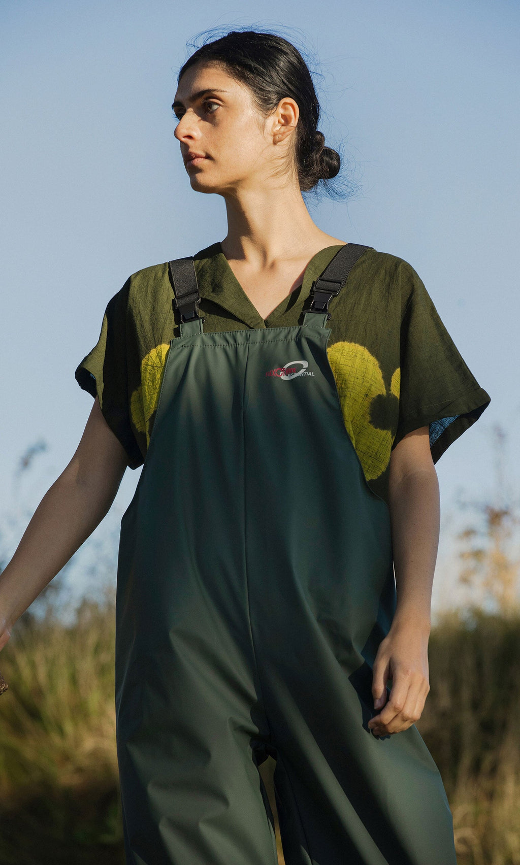 Plümo- A woman stands outdoors under a clear blue sky wearing weatherproof Land Dungarees and a dark patterned shirt.