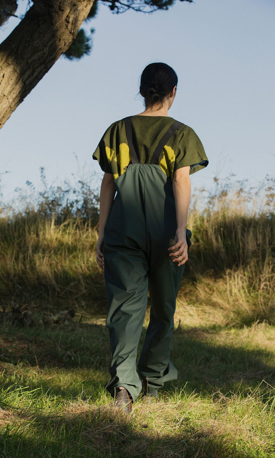 Plümo- Someone in Land Dungarees and a yellow shirt walks uphill on grass beneath a clear sky.