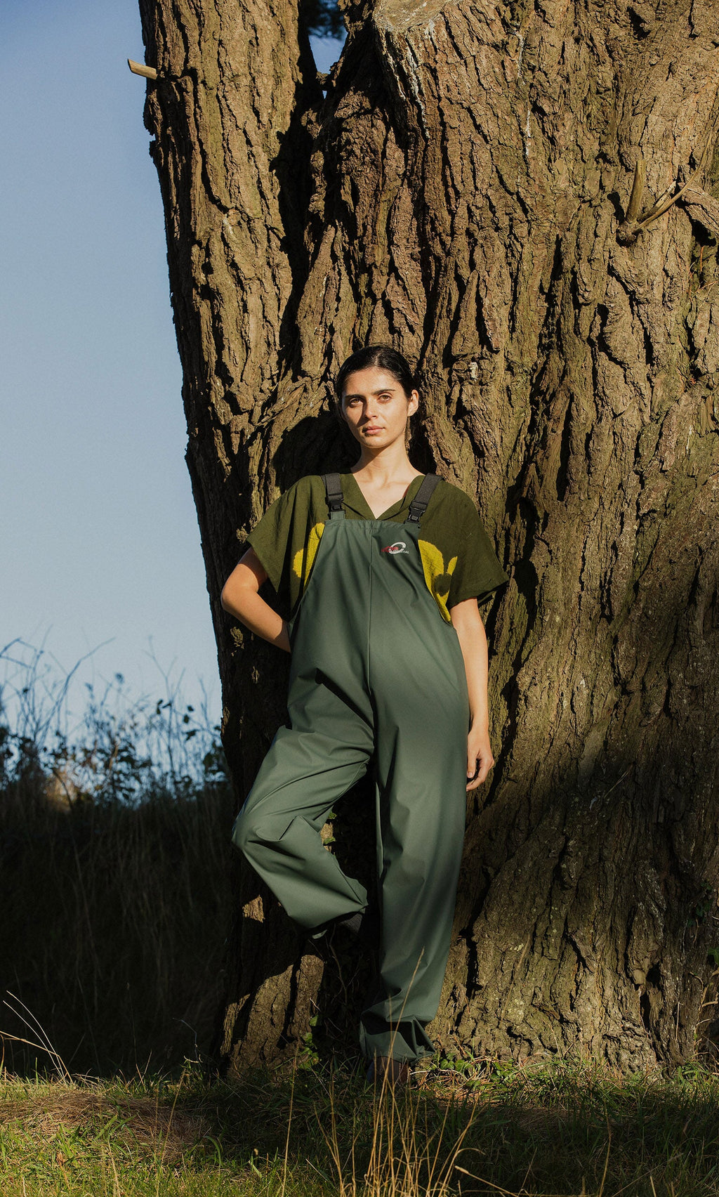 Plümo- A woman wears Land Dungarees and a t-shirt as she stands by a tree trunk outdoors in the sunlight.