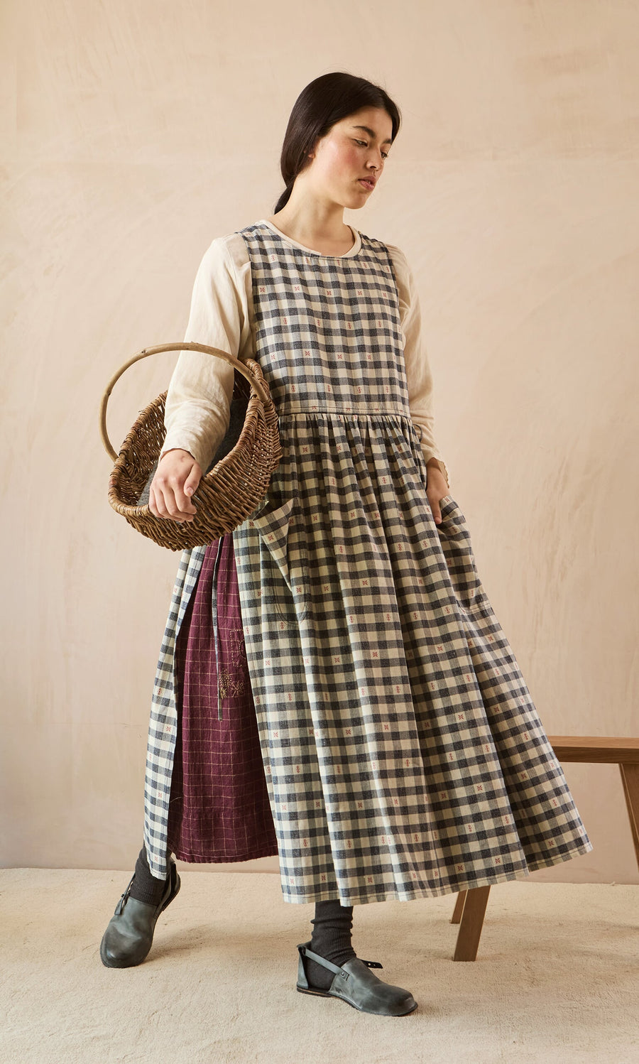 Plümo- Woman in OMalley Apron Dress with adjustable ties & big pockets, over a cream shirt, holding a wicker basket.