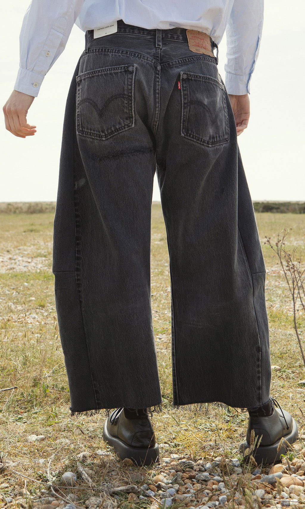 Plümo- Person standing outdoors in Lasso Jeans - Vintage Black, white shirt, and black shoes, viewed from behind.