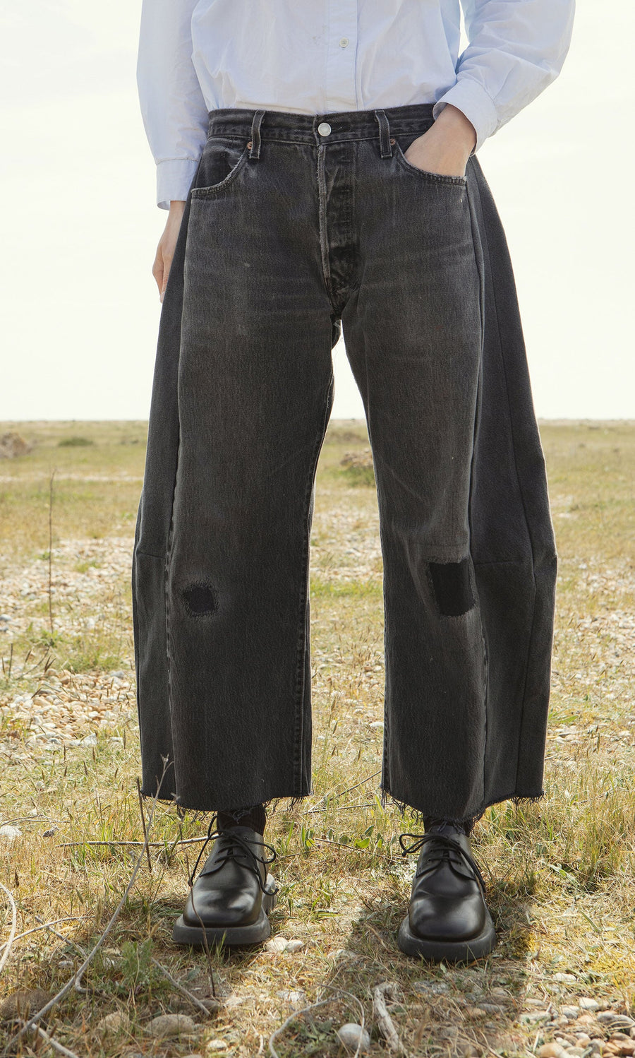 Plümo- Person standing outdoors in Lasso Jeans - Vintage Black, black shoes, light shirt; hands in pockets.