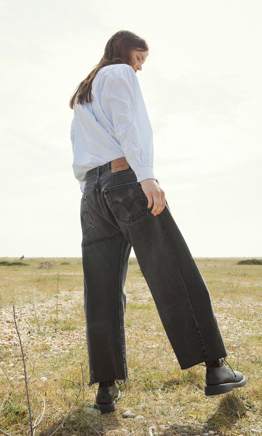 Plümo- Person in a light blue shirt and Lasso Jeans - Vintage Black stands on grass, looking down with one foot slightly raised.