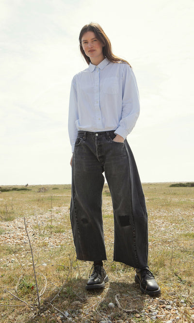 Plümo- A woman stands on grass in a light blue shirt and Lasso Jeans – Vintage Black, hands casually in her pockets.