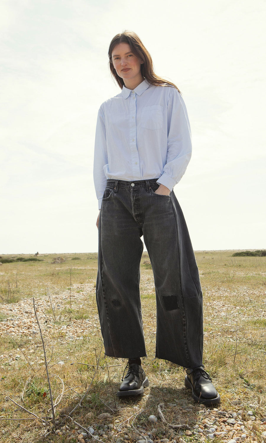 Plümo- A woman stands on grass in a light blue shirt and Lasso Jeans – Vintage Black, hands casually in her pockets.