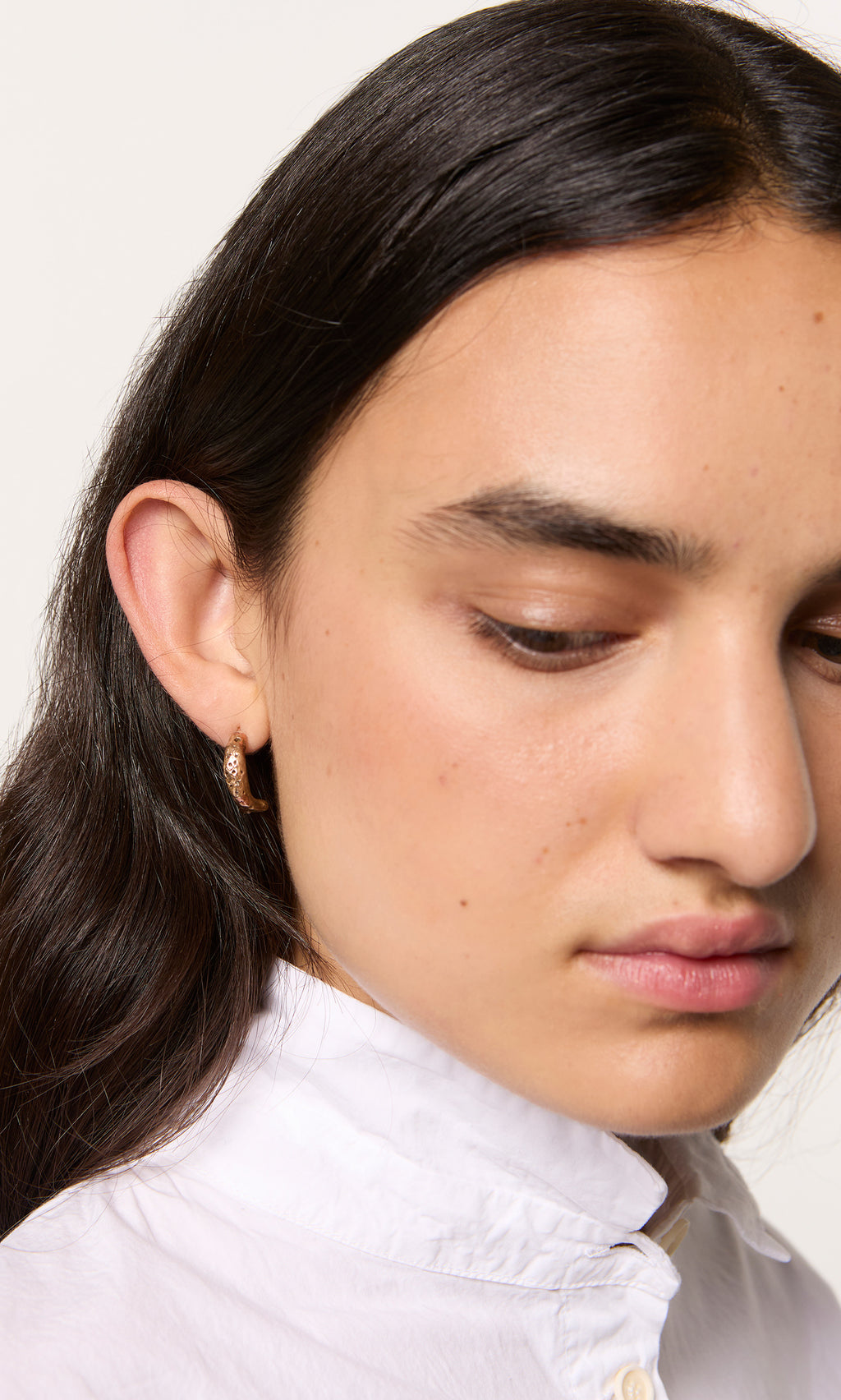 Plümo  Person with long dark hair in a white shirt, wearing Leaping Fish Hoops, looks down.