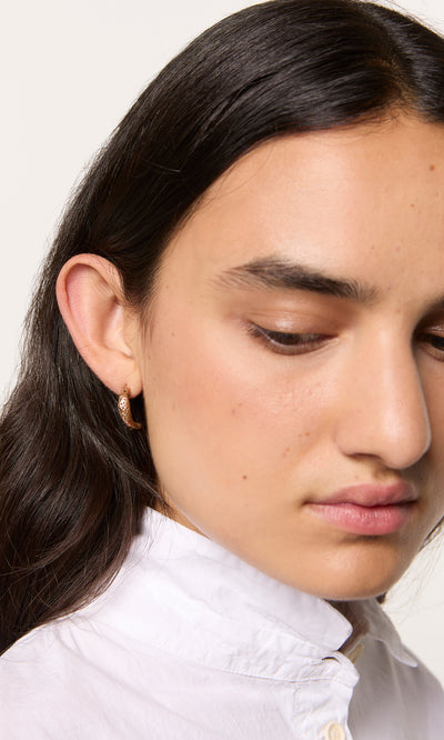 Plümo  Person with long dark hair in a white shirt, wearing Leaping Fish Hoops, looks down.