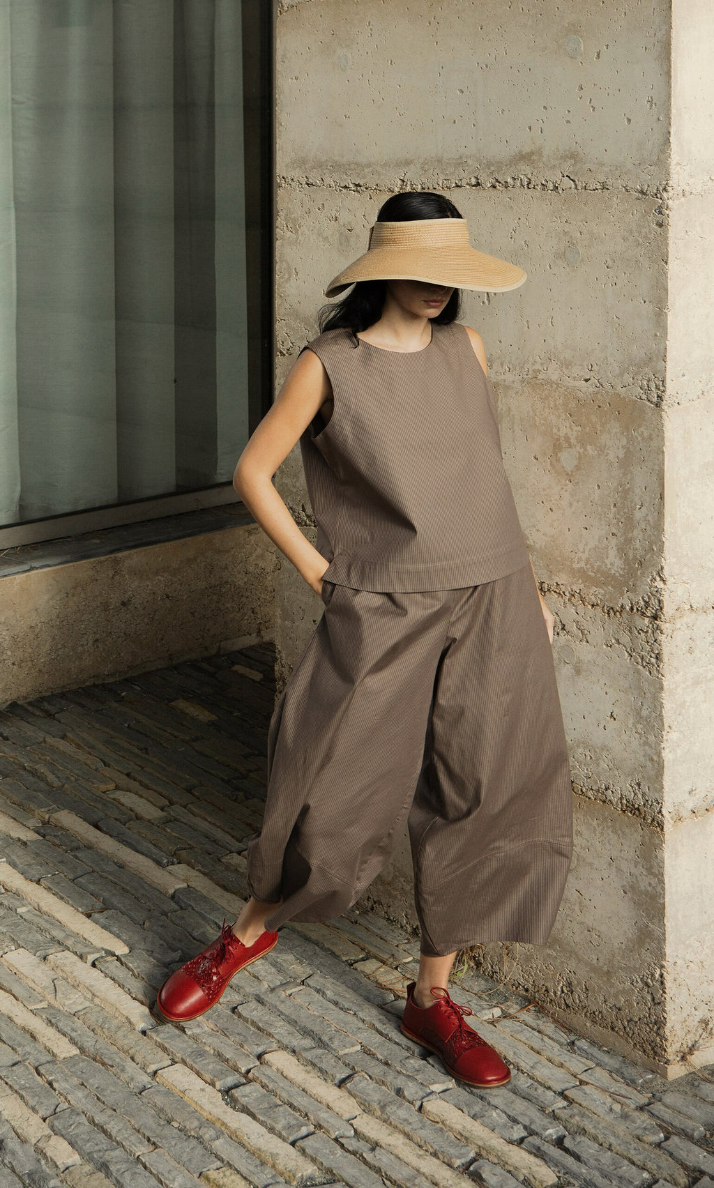 Plümo- A woman in a Linda Top - Brown Stripe, brown outfit, and red shoes stands by a stone wall on a cobblestone path.
