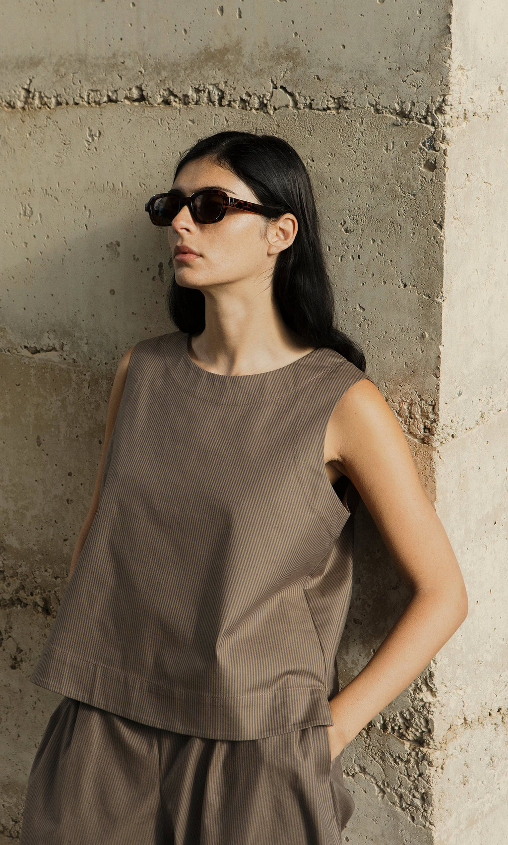 Plümo- Woman with long black hair in sunglasses leans on a wall, wearing the Linda Top - Brown Stripe and matching pants.