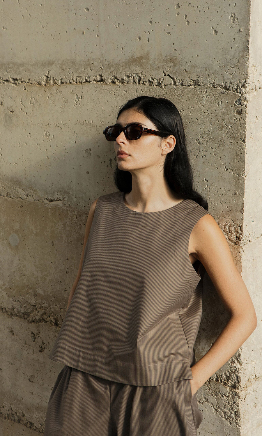 Plümo- Woman in Portia Sunglasses and a brown sleeveless outfit leans against a textured concrete wall, looking to the side.