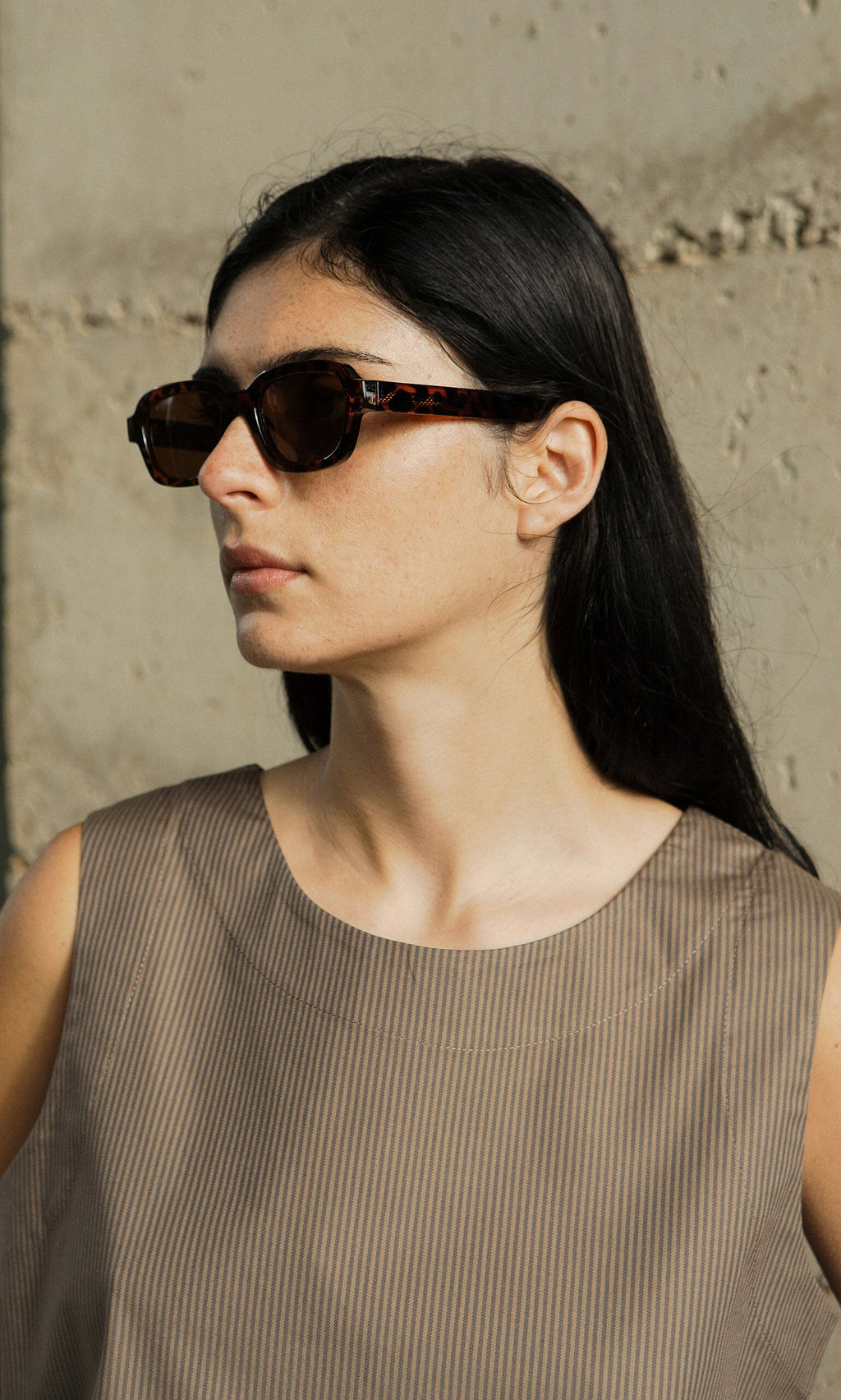Plümo- Woman with long dark hair wearing Portia Sunglasses and a sleeveless taupe top, standing by a concrete wall.