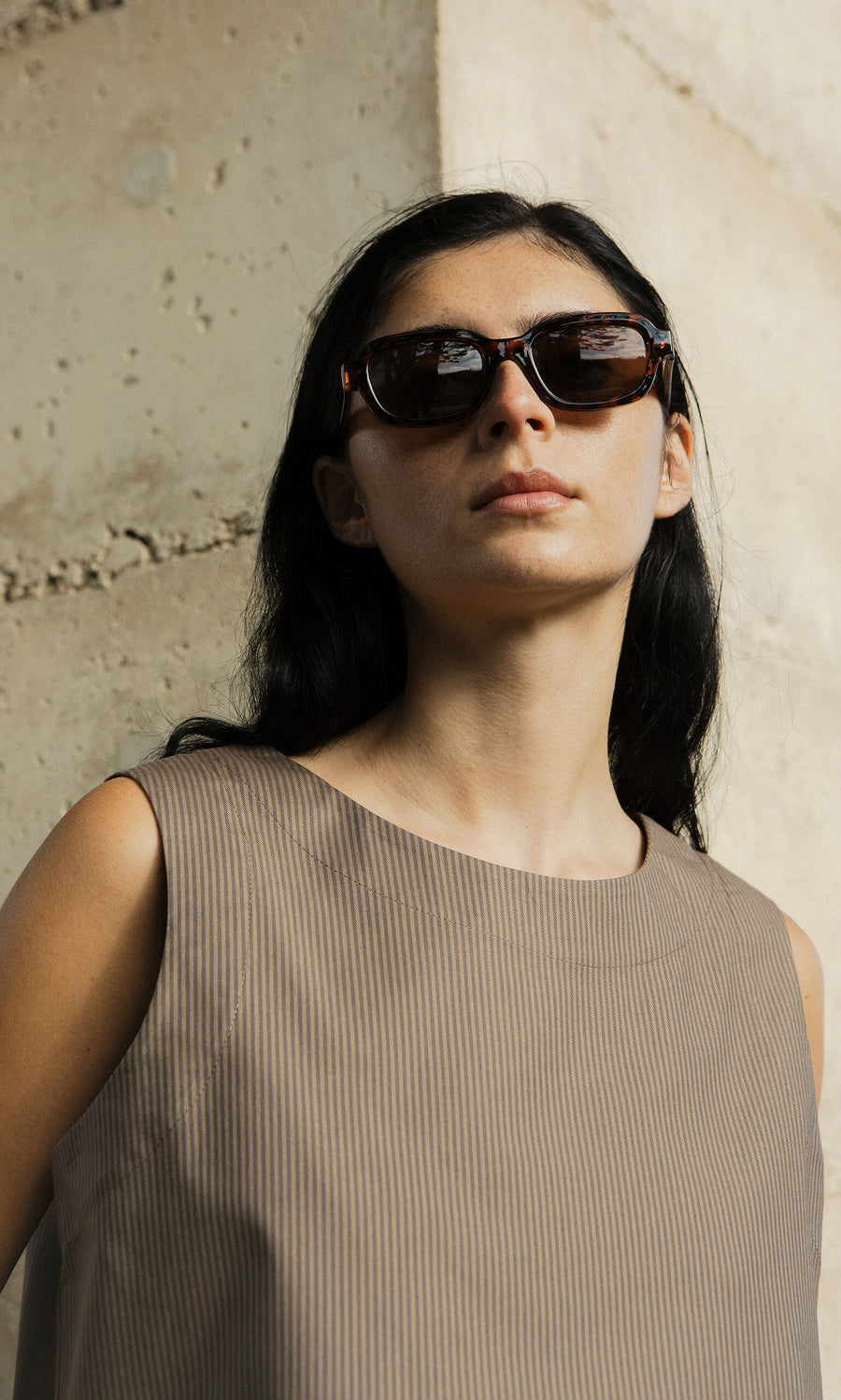 Plümo- Woman with long dark hair wears Portia Sunglasses and a sleeveless beige top, standing by a concrete wall.