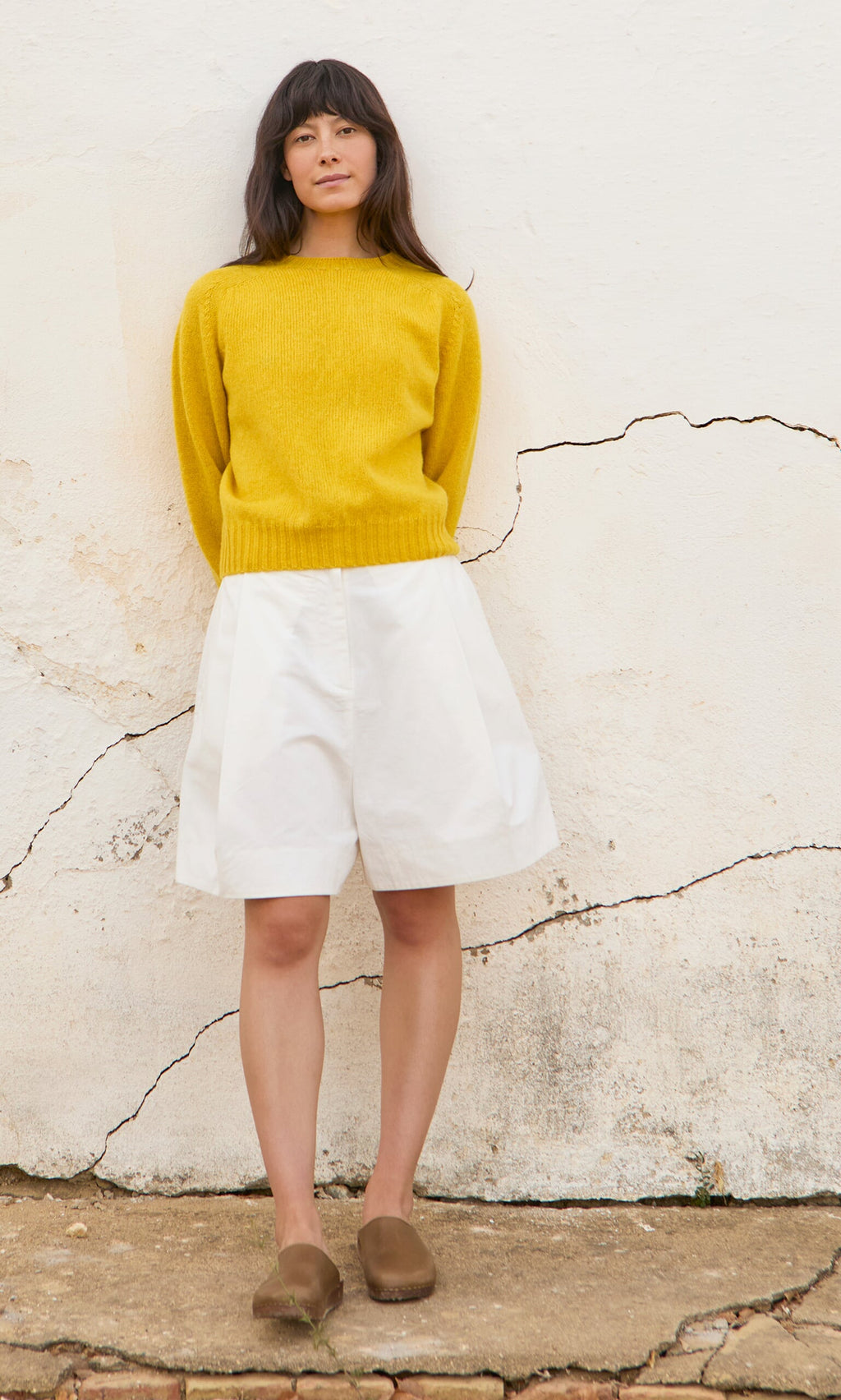 Plümo- Woman in a yellow sweater and Lola Shorts stands against a cracked white wall, wearing brown shoes.