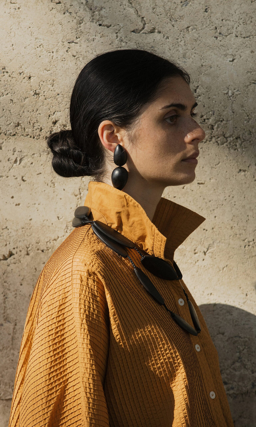 Plümo- Woman in a textured mustard shirt wearing the Benin Necklace and large black earrings, standing by a stone wall in sunlight.