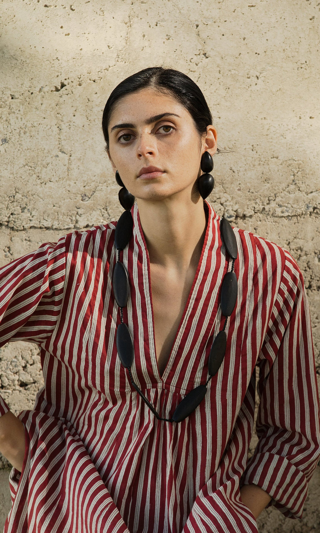 Plümo- Woman in a red and white striped dress wears the Benin Necklace, standing by a textured beige wall.