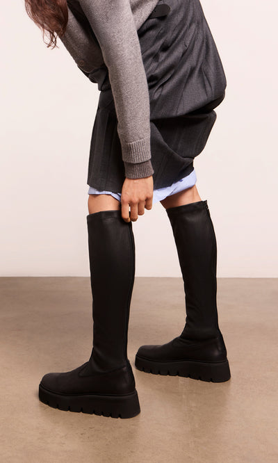 Plümo  Person in Long Prato Boots with chunky soles, gray skirt, and cardigan, bending on a beige floor.