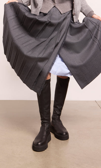 Plümo  Person in Long Prato Boots, black with chunky soles, gray pleated skirt lifted to reveal a light blue underskirt.