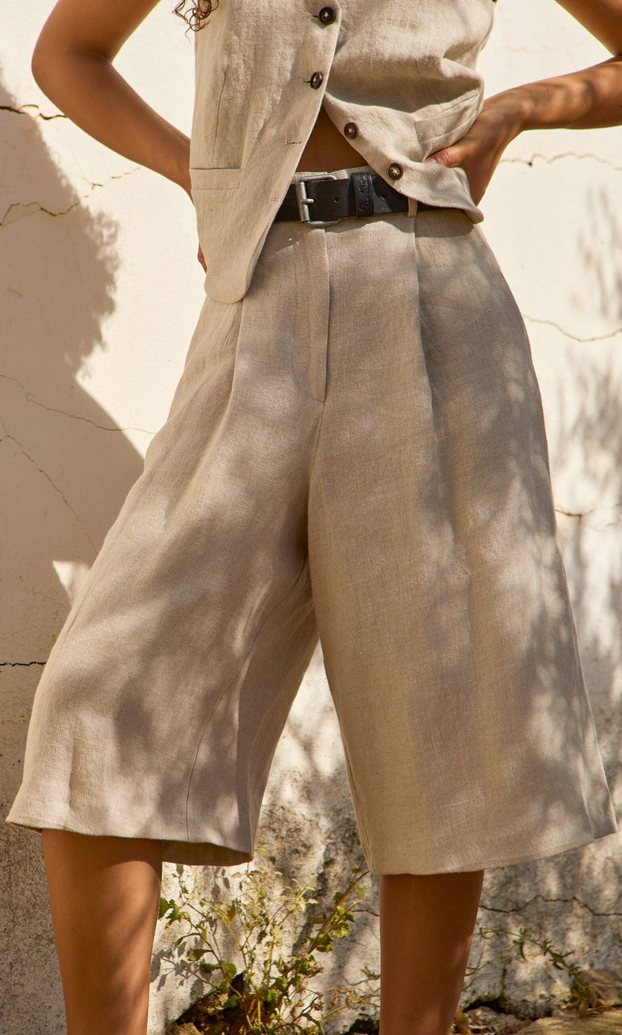 Plümo- Wearing Lucien Shorts - Natural, a person stands with hands on hips against a cracked, sunlit wall.