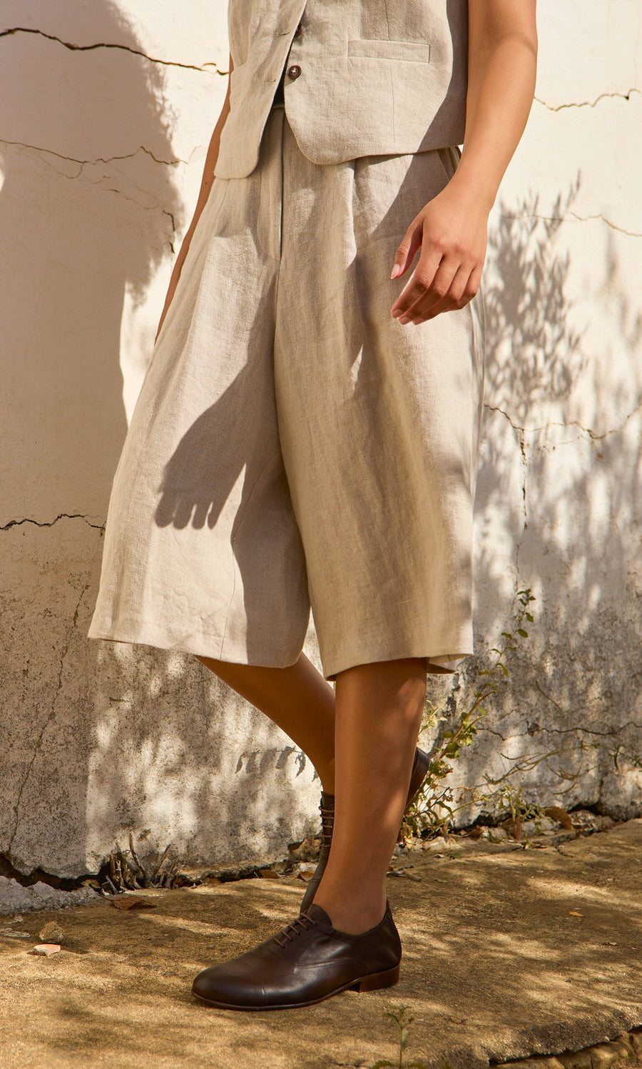 Plümo- Standing outdoors in Lucien Shorts - Natural, a matching vest, and dark brown lace-ups against a textured wall.