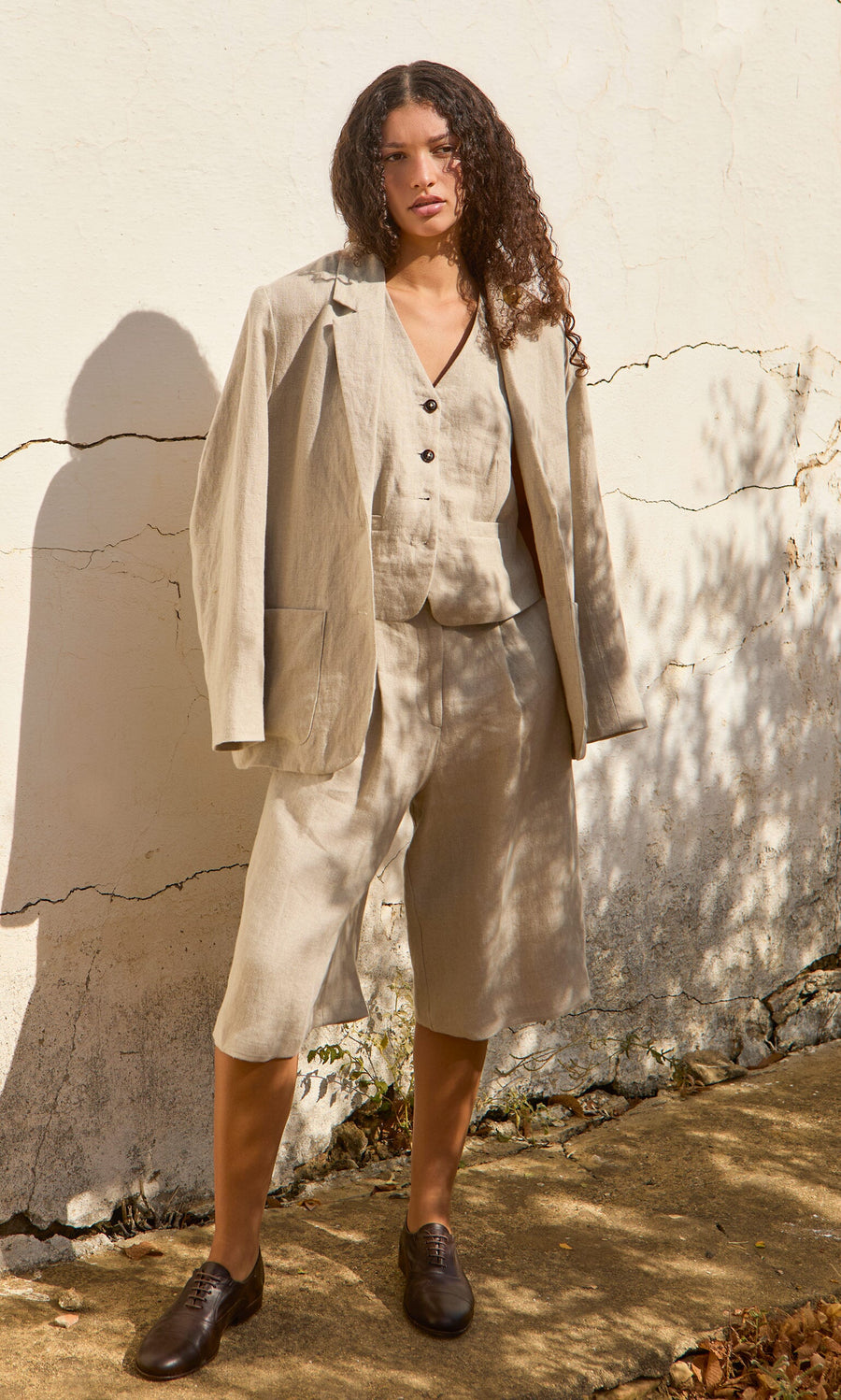 Plümo- Woman outdoors in Lucien Shorts - Natural, a beige Italian linen Bermuda shorts suit, paired with brown shoes.