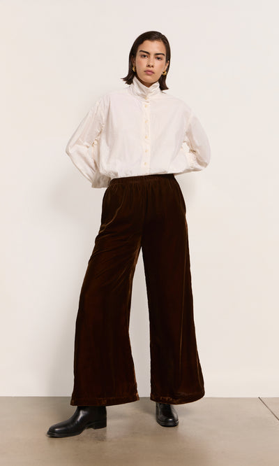 Plümo  Person in a white button-up shirt, Maroon Velvet Pants with wide legs, and black boots.