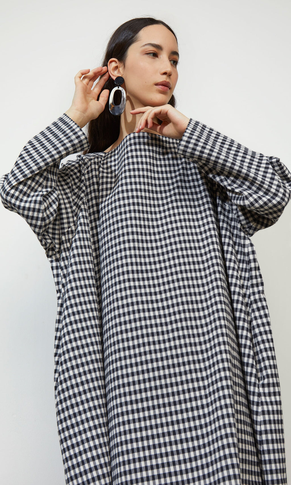 Plümo- Woman wearing the Marsell Dress - Gingham, holding one hand near her ear and looking to the side.