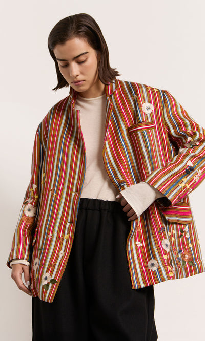 Plümo  Person in a silk blazer with colourful red stripes and hand-stitched flowers, cream shirt, black pants, plain background.